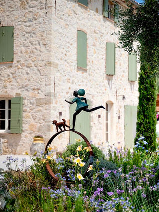 Bronze sculpture of a girl in a green dress running across the top of a curved metal hoop, holding a lead attached to a small dog, set among a colourful meadow of blue, purple and yellow flowers. A stone country house with pale green shutters forms the backdrop.