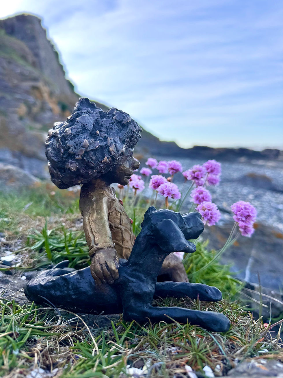 Thumbnail: Bronze child and black dog sculpture nestled among sea thrift flowers on rocky ground, child in quilted red-button top.