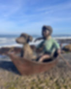 Bronze boy rowing a boat with his dog, wearing a textured mint jumper and scarf, among rocks beside the sweeping coastline.