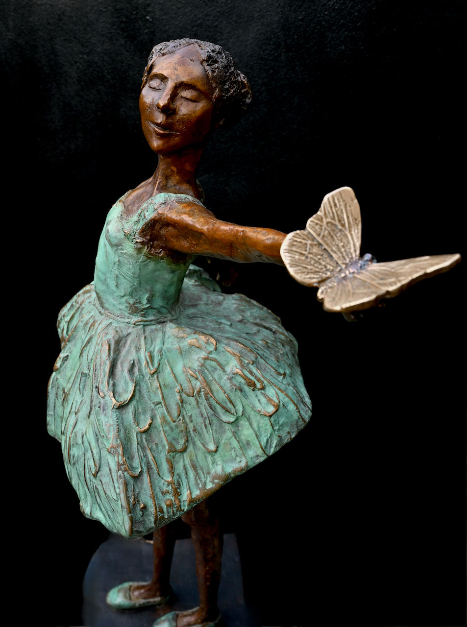 Thumbnail: Close view of ballet sculpture’s feather-textured skirt and serene face as she lifts a butterfly in one hand.
