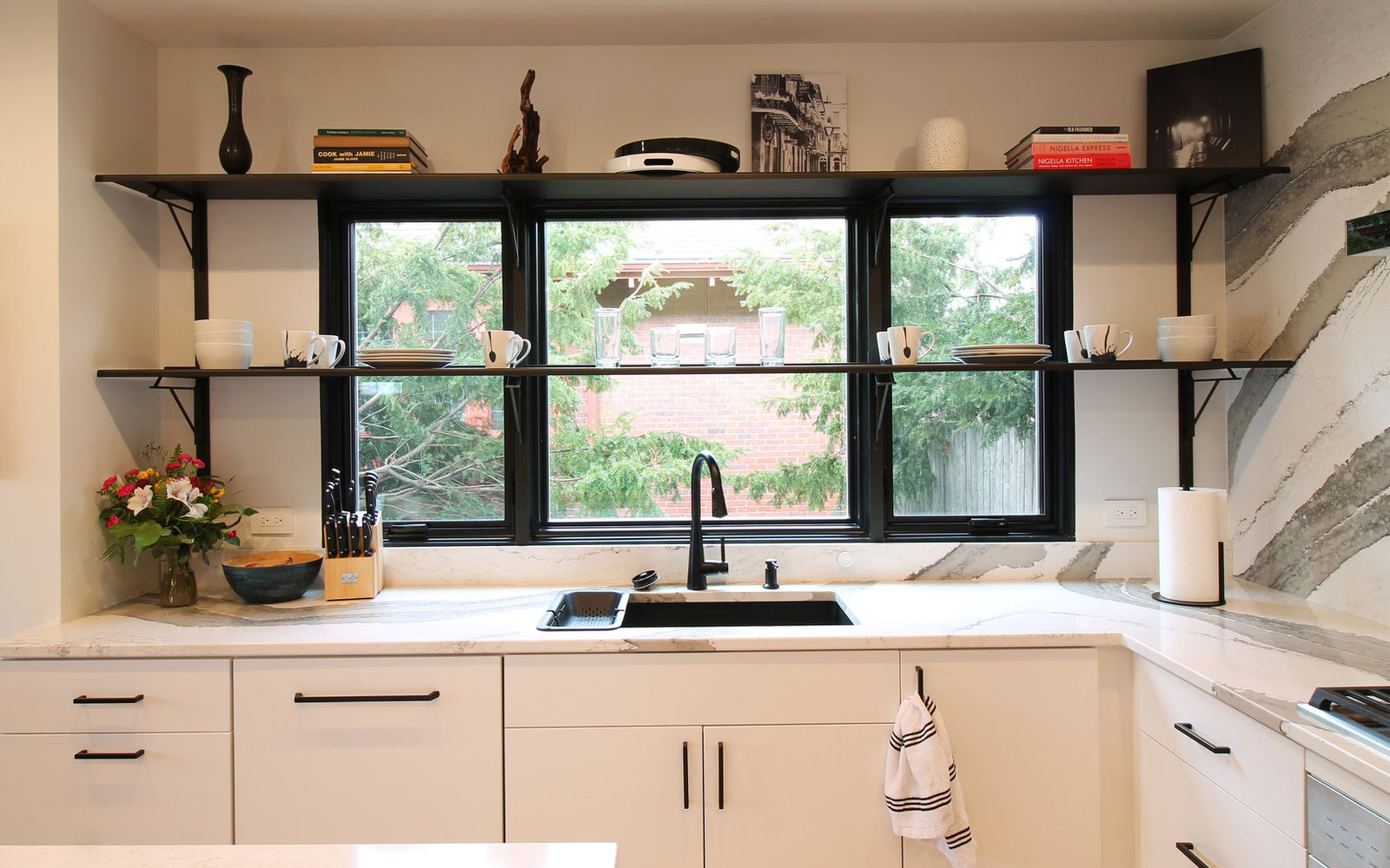 Modern kitchen with white cabinets, black faucet, and large window showing greenery. Shelves hold books, cups, and decor. Bright and tidy.