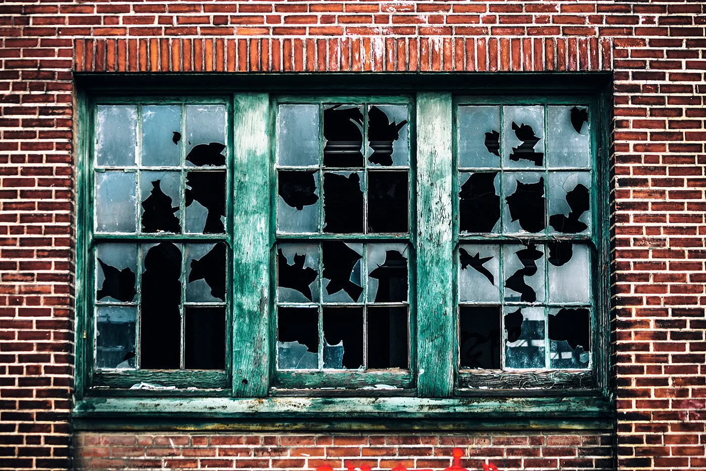 Old brick building with a green-framed window, panes shattered and missing. Weathered texture, graffiti at the bottom, evokes abandonment.