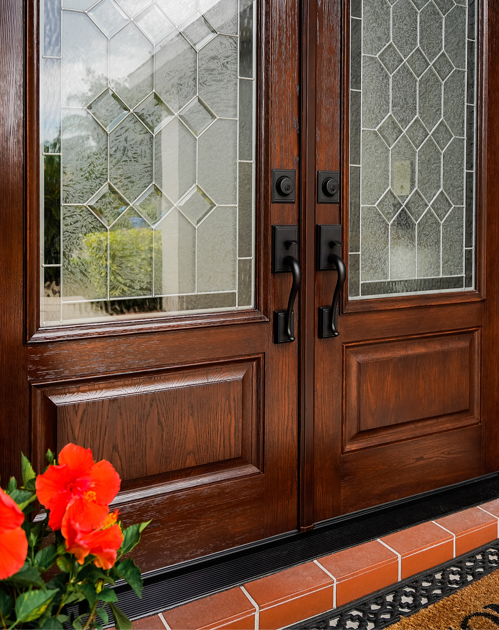 Wooden double front door with frosted stained-glass panels and black handles, beside bright orange flowers on a brick stoop.