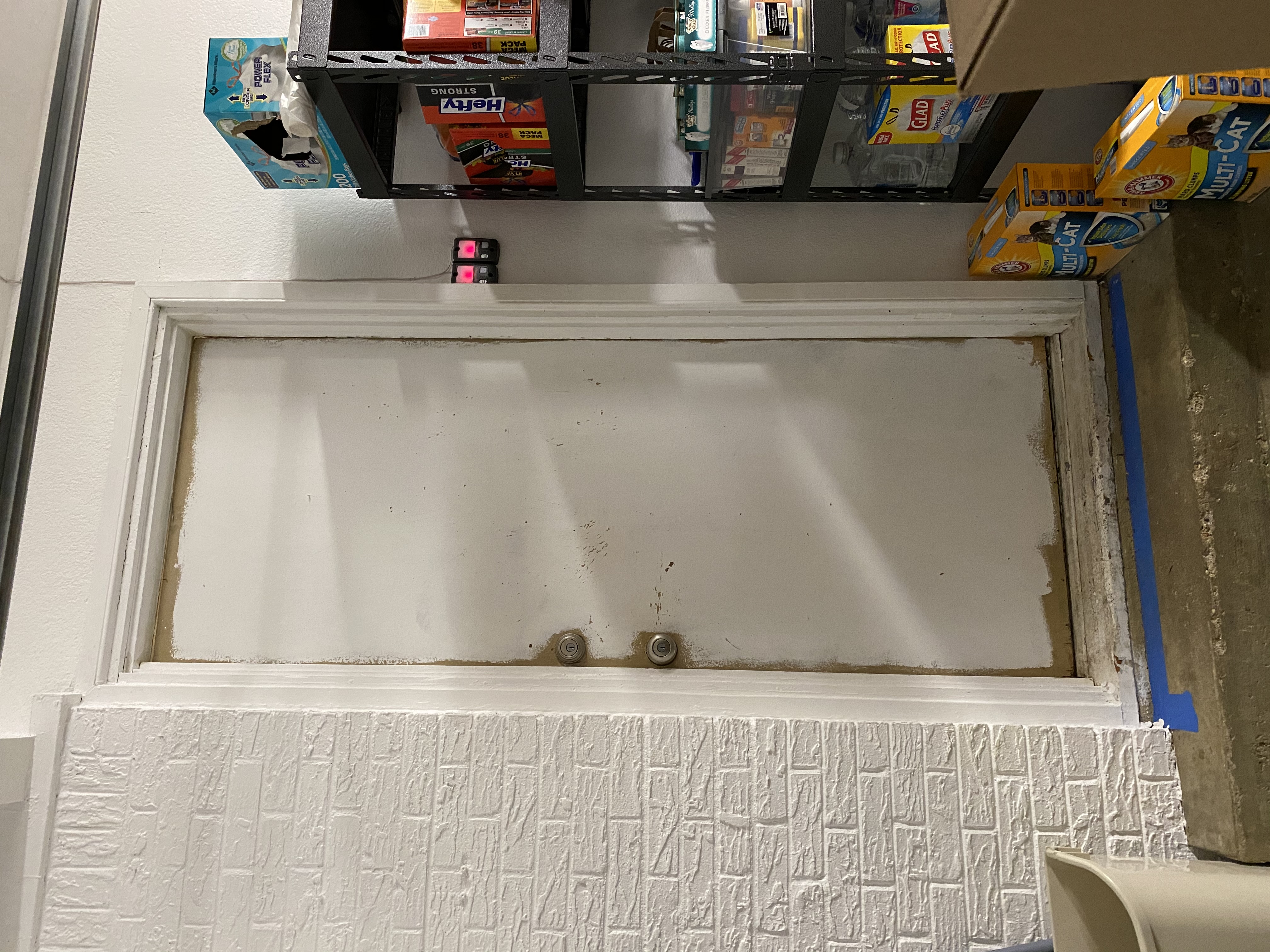 White door with two knobs in a brick wall, surrounded by a shelf with cat litter and cleaning supplies. Dimly lit interior setting.