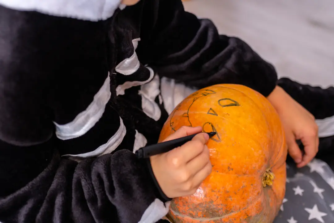 A kid dressed in a Halloween costume drawing a face on a pumpkin.