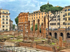Ancient ruins with stone columns in foreground, surrounded by colourful historic buildings and a large tree. Overcast sky in background.