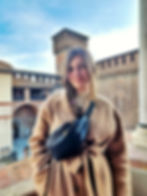 A woman in a beige coat smiles at the camera. She is standing on a balcony of a castle, with the rest of the structure (including a turret) visible behind her. The sky is blue.