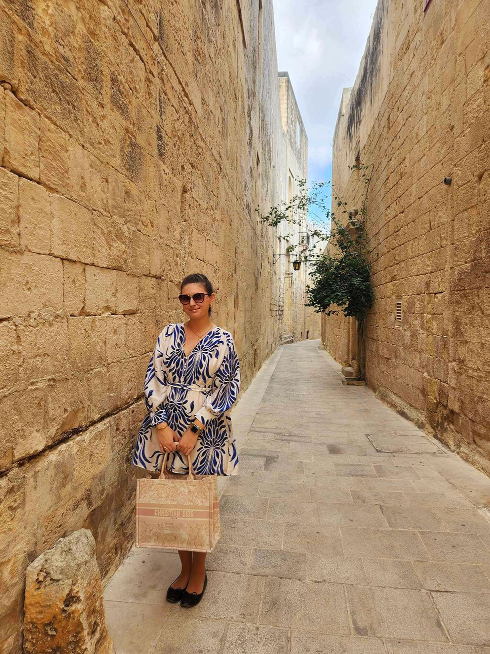 Woman in a blue and white patterned dress holds a Dior bag in a narrow stone alley. Sunny day, relaxed mood, and historic backdrop.