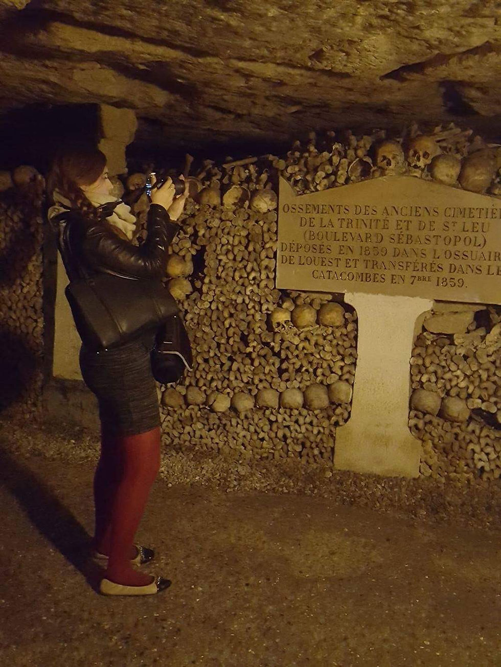A woman photographs skulls and bones in a dimly lit underground catacomb. A plaque with French text is visible, creating an eerie atmosphere.
