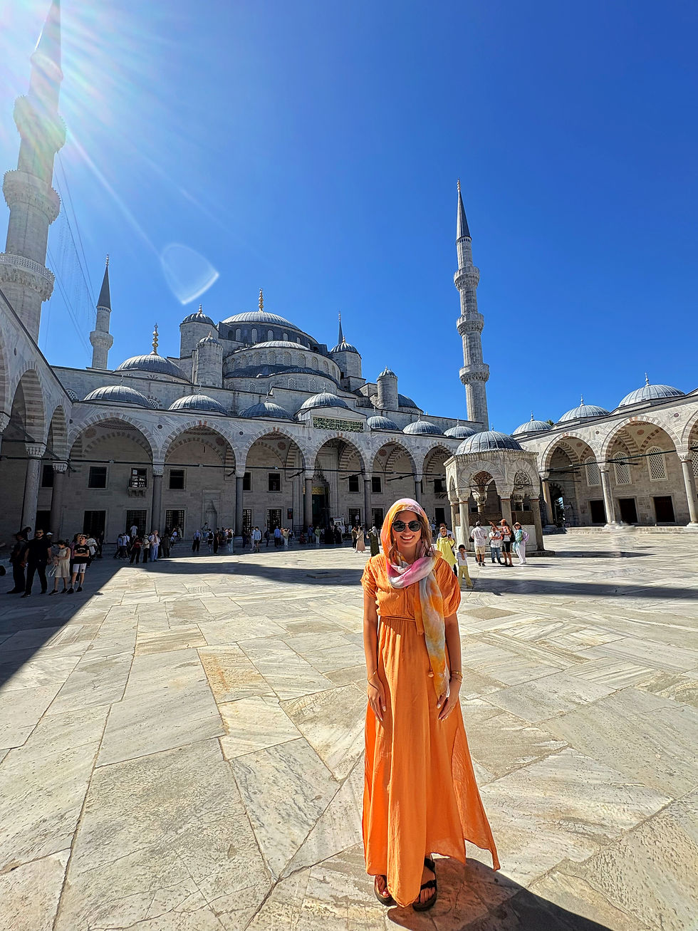 Woman in orange dress and headscarf smiles in a mosque courtyard with domes and minarets under bright blue sky.
