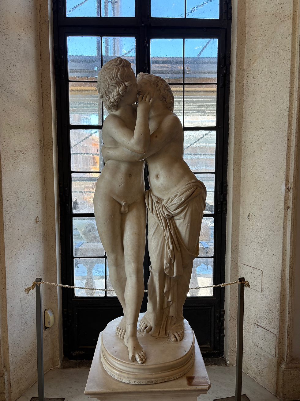 A marble statue of Eros and Psyche embracing in a kiss. The statue is positioned in front of a window.