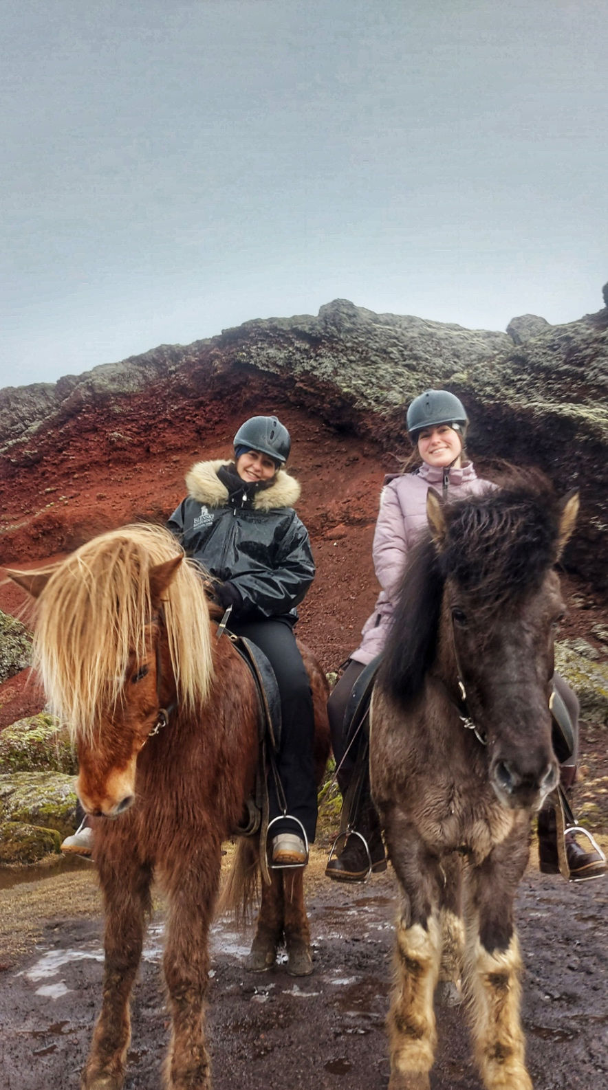 Two women are sitting on horseback, the horses side-by-side, and are smiling at the camera. Behind them are red iron deposits in the volcanic lava of the landscape behind them.