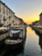 Boats line a canal, the sun setting in a mix of orange and yellow in the sky. There are a number of restaurants with outdoor patios on either side of the canal.