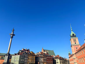 Colorful historic buildings and a statue-topped column under a clear blue sky in a European city, evoking a calm, sunny day.