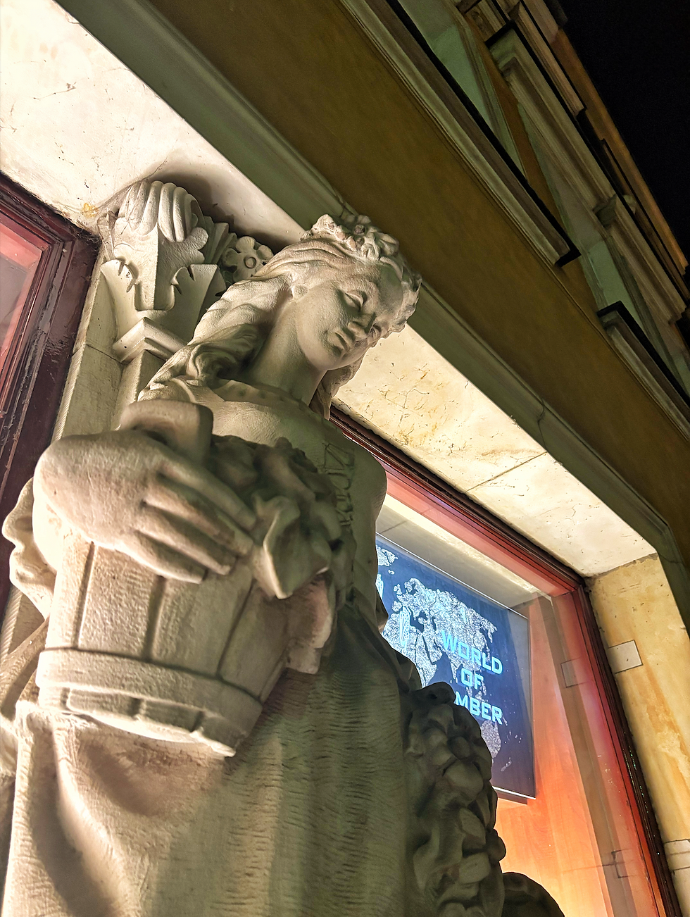 Stone statue of a woman holding a basket, set against a building facade at night. Sign in background reads "World of Amber."