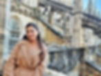 A woman in a beige coat smiles at the camera. Behind her, you can see the intricate designs of Milan's Duomo; she is on the visitors' path of the walkable roof.