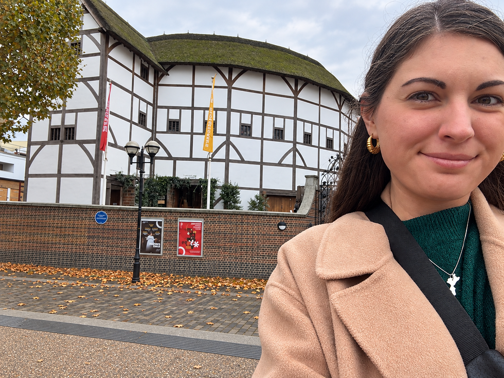 Selfie di una donna sorridente con un cappotto beige davanti a un teatro bianco con struttura in legno, tetto coperto di muschio e foglie autunnali.