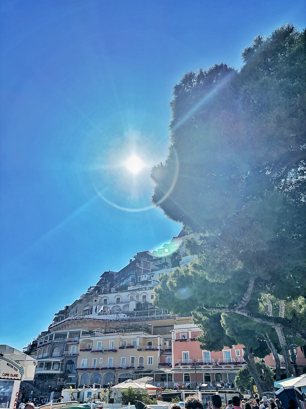 Sunlit buildings on a hillside under a clear blue sky, with a large tree on the right. Sign reading "Capri Island" visible below.