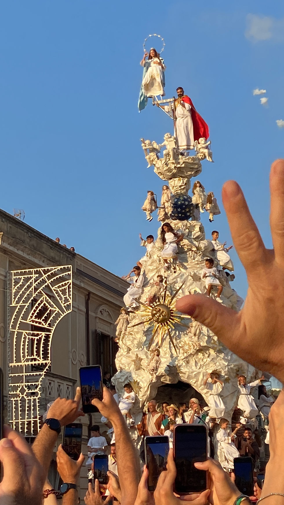 A tall, conical structure adorned with children passes on the street, with a little girl dressed up as the Virgin Mary at the very top.