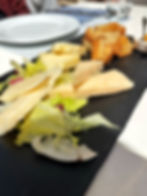 Cheese platter with sliced cheese, green lettuce, and toasted bread on a black slate. Sunlit table setting with empty plates in the background.