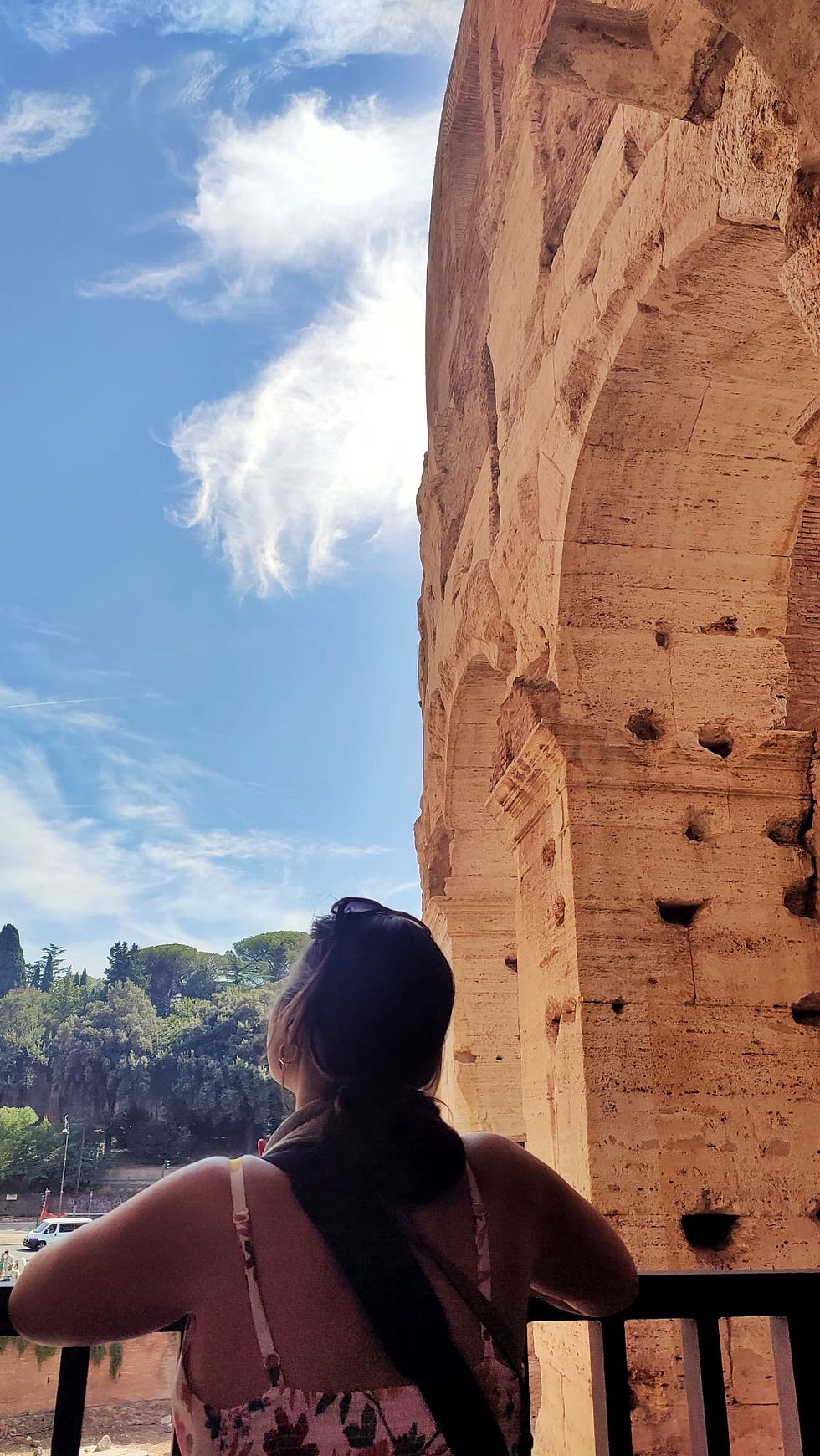 A woman has her back to the camera and is looking up at the stone walls of the Colosseum. In the back is a blue sky with wispy clouds.