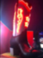 A woman sits on the toe of a giant cowboy boot. The leg of the boot is lit up in a red neon light design.