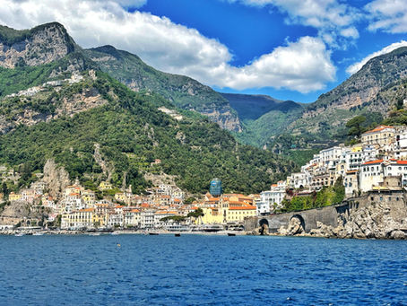 Coastal town with colorful buildings clings to a lush mountainside under a blue sky with clouds. Calm sea in the foreground.