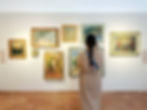 A woman in a beige coat stands in front of a collection of different framed pieces of art in different sizes, hanging on a wall. Her back is to the camera as she looks at the art.