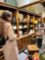 A woman in a store smiles in the reflection of a mirror, handbags on wooden shelves around her. She holds a brown bag. Shelves display colourful bags.