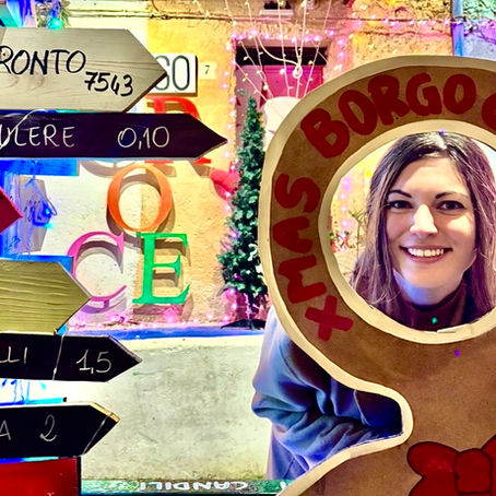 Woman smiles through a gingerbread cutout labeled "BORGO CROCE XMAS." Coluorful signposts point directions, including Toronto. Festive lights create a joyful ambiance.