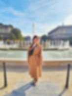 A woman in a beige coat smiles at the camera. She is standing in front of a large fountain spurting water.