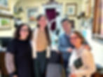 Four people smiling in a warmly lit room filled with framed artwork and certificates. One wears a leopard print sleeve sweater. Cozy atmosphere.