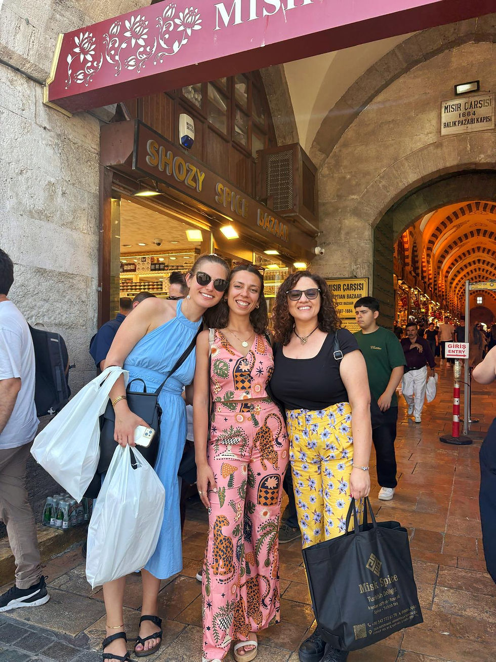 Three smiling women pose outside Istanbul’s Mısır Çarşısı spice bazaar, holding shopping bags.