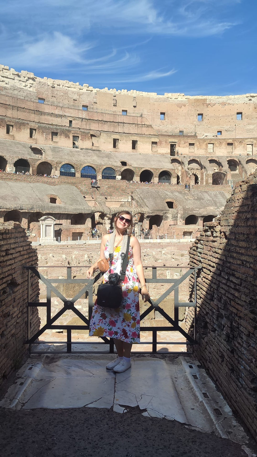 A woman in a floral sundress and a camera around her neck smiles at the camera. In the background is the Roman Colosseum.