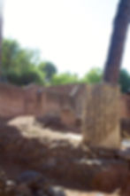 The remains of a carved pillar stands amongst the ruins. In the background are old brick walls still standing.