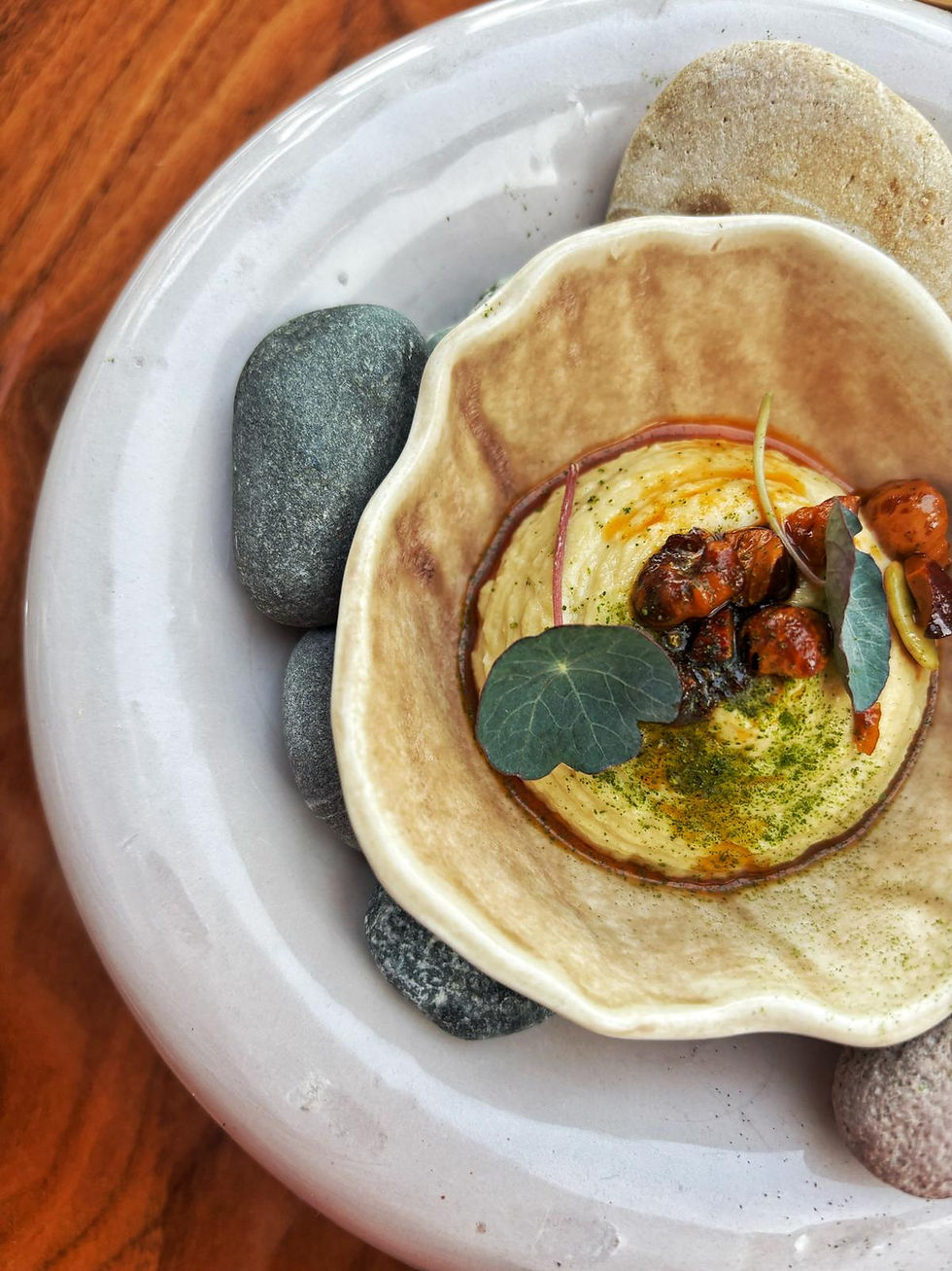 Gourmet hummus-like dish in a ceramic bowl on a white plate with smooth stones, on a wooden table.