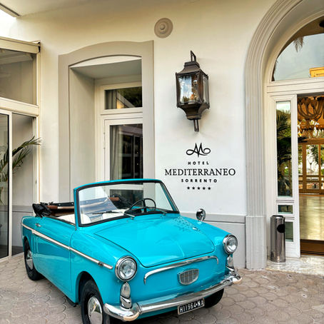 Bright turquoise vintage convertible parked at Hotel Mediterraneo Sorrento entrance, with black wall lantern and hotel sign visible.