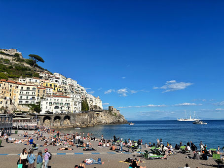 Sunny beach below cliffside town with crowded sunbathers, blue sea, sailboat and yacht under clear sky.