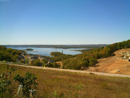 Overlooking a lake with calm blue water surrounded by wooded hills and a small settlement. Clear sky and earthy tones dominate the serene scene.