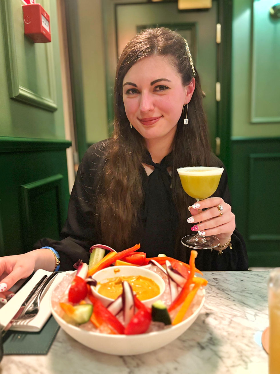 Woman with long hair and a black outfit smiles while holding a yellow cocktail. A colorful vegetable platter is on the marble table. Green wall behind.