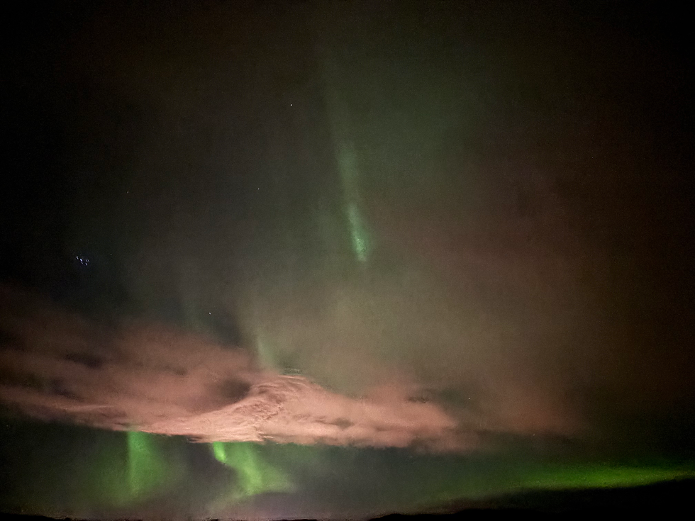The green lights of the Aurora Borealis dance in a dark sky. Illuminated by it are some reddish-coloured clouds.