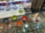 Colourful jewelry shop display with painted ceramic bowls, gold necklaces, a calculator, and glass cases filled with gemstone jewelry.