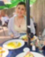 A woman sits at a table smiling. In front of her, on the table, are a variety of delicious-looking breakfast items.