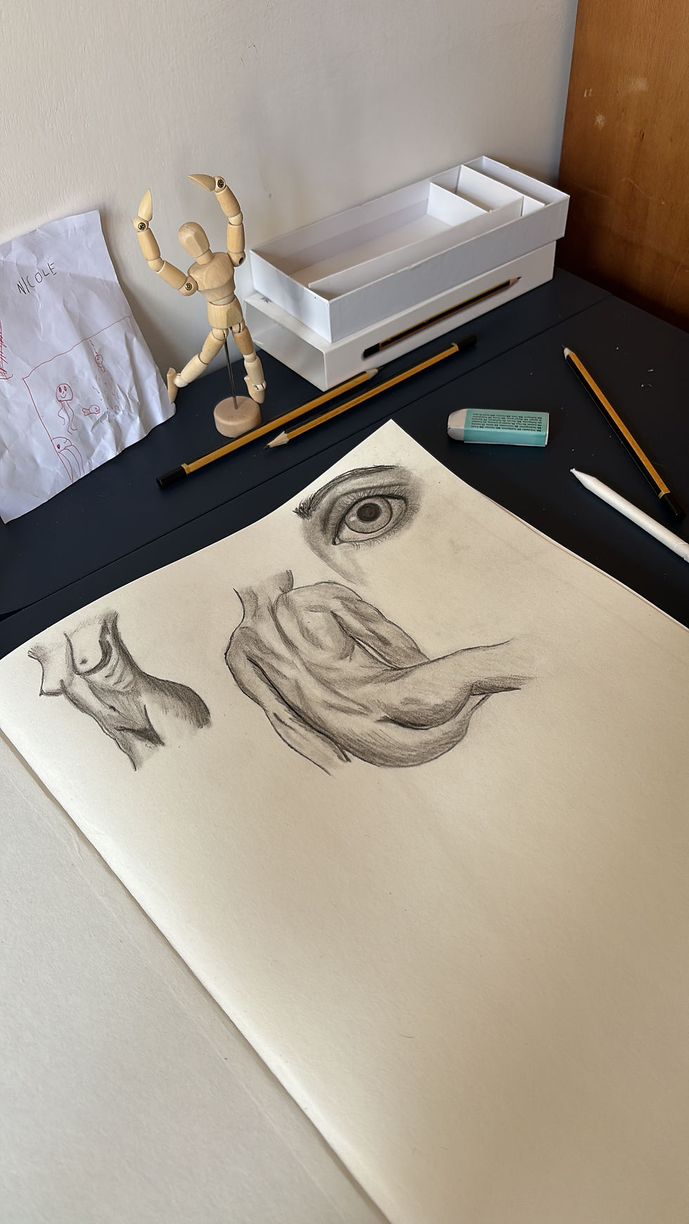 Sketchbook with eye and anatomy sketches, surrounded by pencils, eraser, a wooden mannequin, and a child's drawing on a desk.