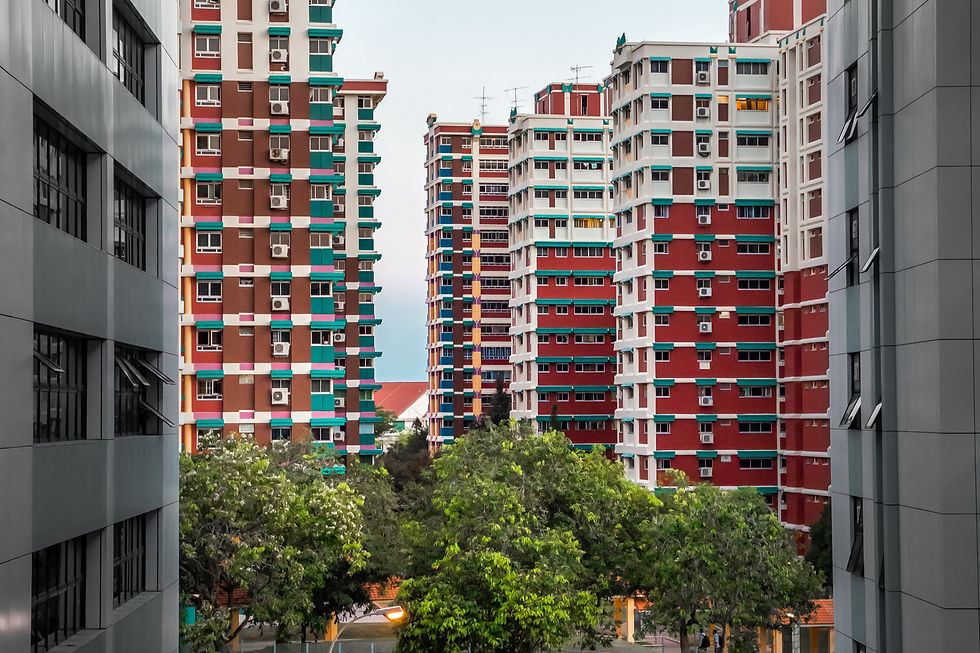 Colorful high-rise buildings in a cityscape with red and green accents. Trees in the foreground, cloudy sky in the background.