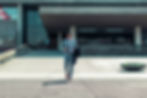 Man in a gray suit walking outside a modern building, checking his phone. Holds a dark coat. Sunny day with a flower pot nearby.