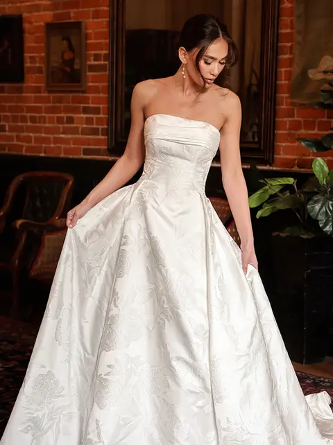 Designed for the modern romantic, Style D4304 is a stunning satin ballgown that blends timeless elegance with contemporary charm. The strapless, straight-across neckline folds over a structured bodice, creating a refined yet effortlessly chic silhouette. Subtle seaming contours the figure before meeting the voluminous pleated skirt at the hips, delivering a regal statement. In the back, a row of fabric-covered buttons traces the length of the gown, adding a classic touch. With its grand yet minimalist appeal, this wedding dress is the ideal choice for the fashion-forward bride seeking sophistication with a hint of drama. This gown is also available in plus sizes.
