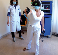 Une séance de rééducation en réalité virtuelle médicale est en cours dans un hôpital. Une patiente avec des manettes de RV bouge le bras droit avec un casque VR sur la tête. Au fond de la salle de rééducation, sa kinésithérapeute la regarde. À côté de la kiné, se trouve le dispositif de rééducation en réalité virtuelle KineQuantum dans une armoire médicale.