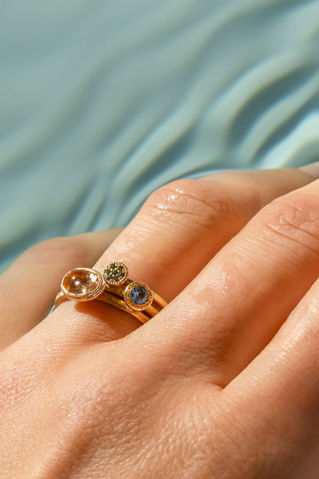 Drei Ringe aus recyceltem Gold mit fair gehandelten Edelsteinen auf einer Hand vor Wasseroberfläche.
Michaela Römer sustainable fine jewelry