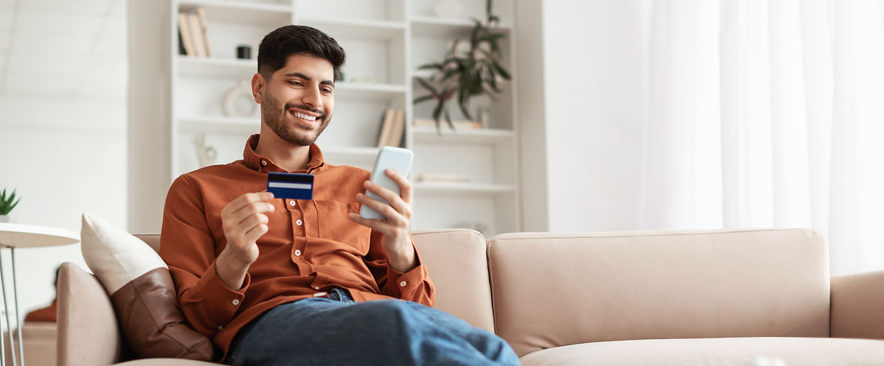 Fast Online Shopping. Smiling young Arab guy holding debit credit card in hand and using c